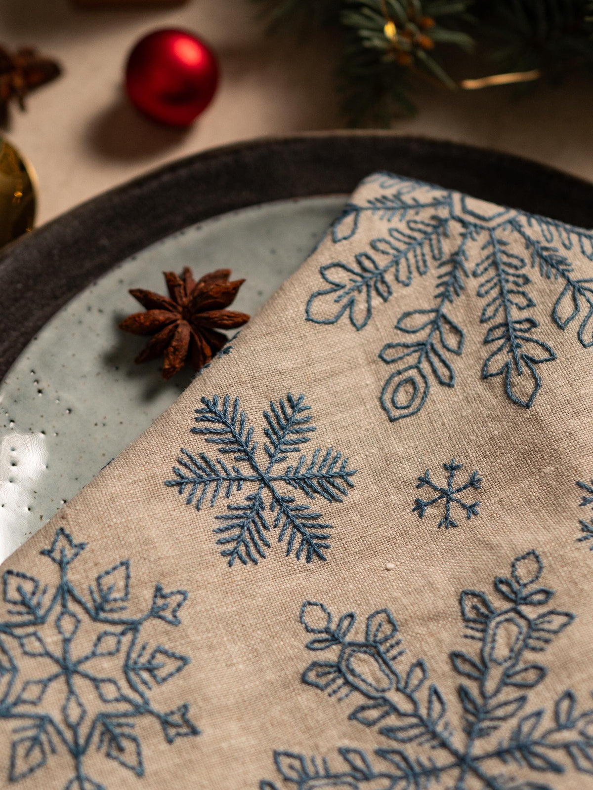 May include: A beige linen napkin with embroidered blue snowflakes, folded on a light blue plate. The napkin features various snowflake designs. A red Christmas ornament and star anise are visible in the background, suggesting a holiday theme.