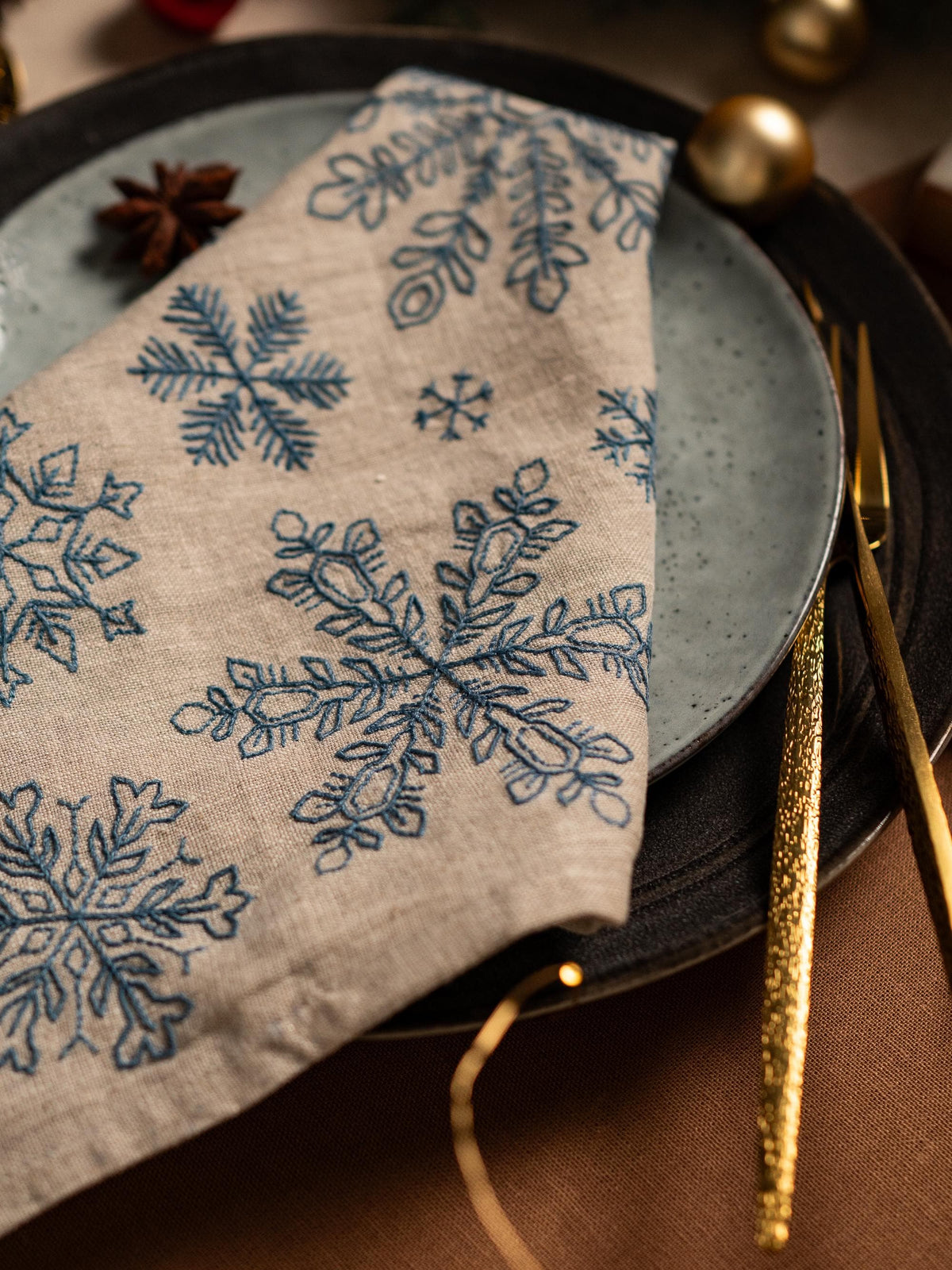 May include: A beige linen napkin with embroidered blue snowflakes rests on a plate. The napkin is folded, displaying several snowflake designs. A gold fork and knife are next to the plate, suggesting a festive dining setting. The plate is a muted bl