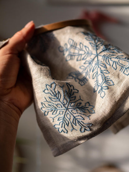 May include: A linen fabric with blue embroidered snowflake designs. The fabric is held in a wooden embroidery hoop. The embroidery thread is a deep blue color, contrasting with the natural beige fabric. The design features detailed snowflake pattern