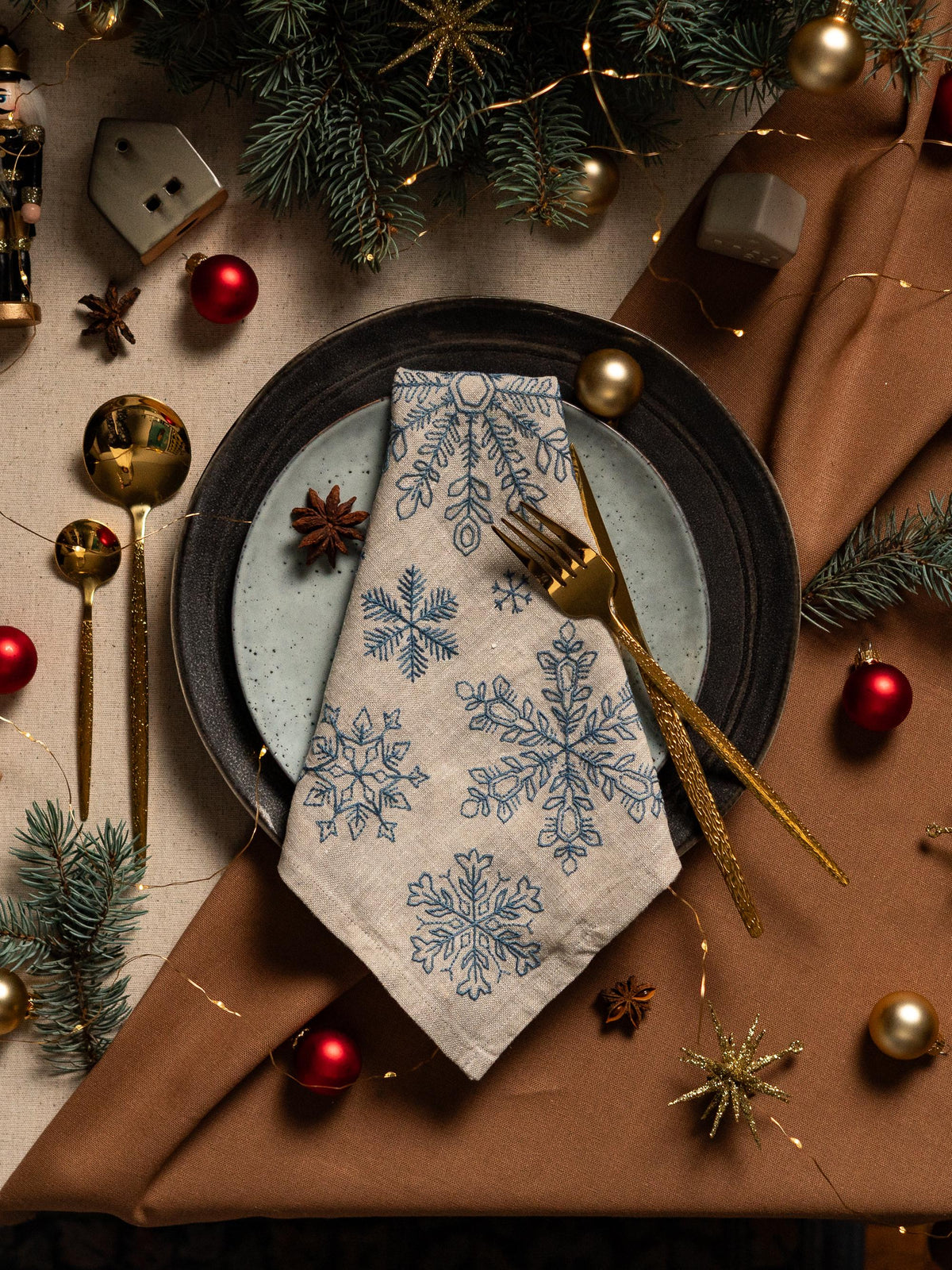 May include: A festive holiday table setting with a linen napkin featuring blue snowflake embroidery. Gold-colored flatware, red and gold ornaments, and pine branches complete the seasonal decor. A black plate and a light blue plate are also visible.