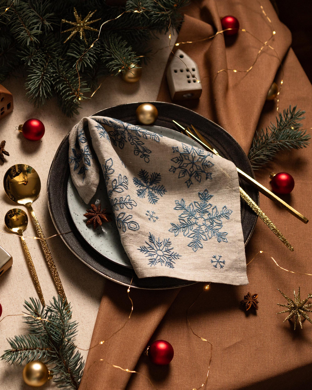May include: A festive table setting with a linen napkin featuring blue snowflake designs. The plate is set with gold utensils, red ornaments, and pine sprigs. The overall theme is Christmas or winter holiday decor.