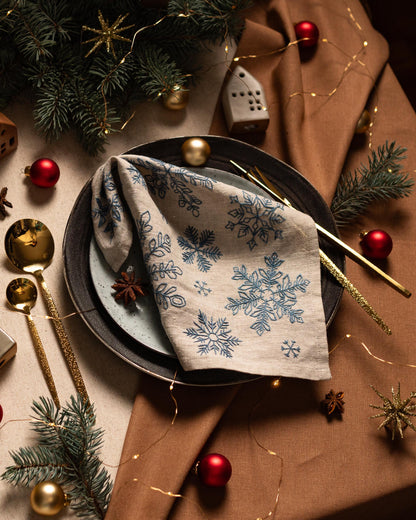 May include: A festive table setting with a linen napkin featuring blue snowflake designs. The plate is set with gold utensils, red ornaments, and pine sprigs. The overall theme is Christmas or winter holiday decor.