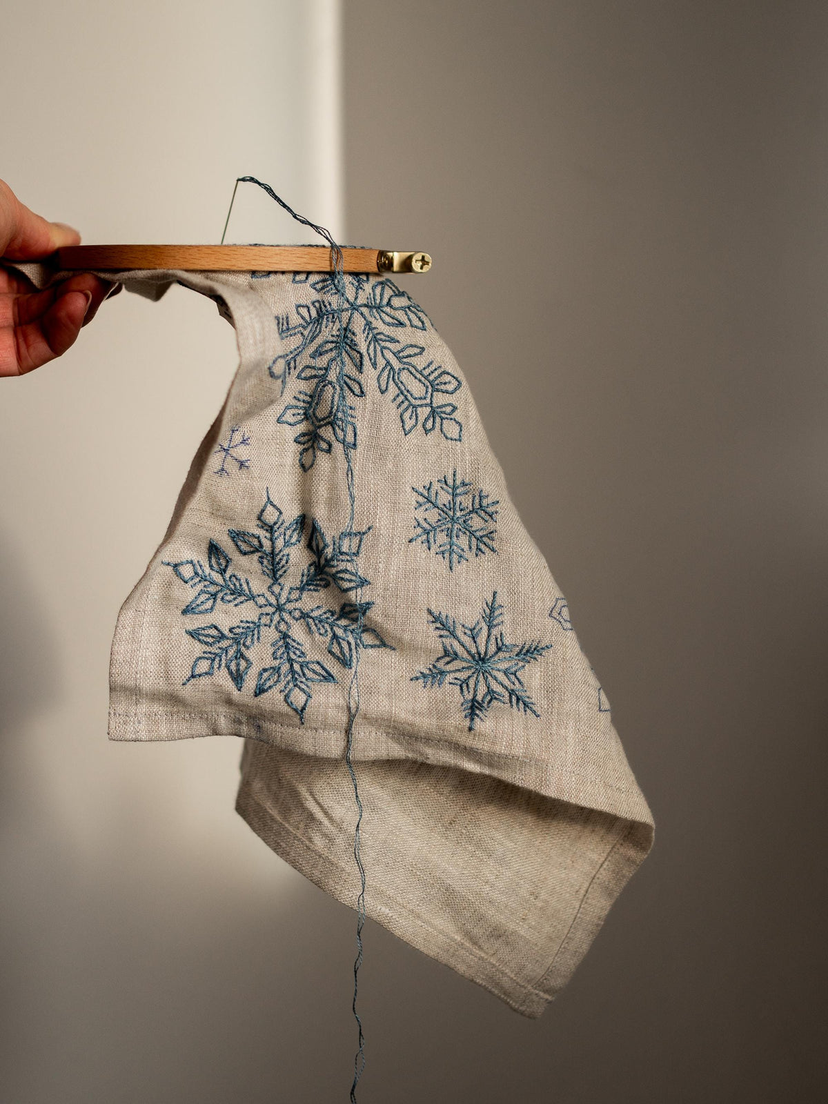 May include: A linen fabric with blue snowflake embroidery is held in a wooden embroidery hoop. The snowflakes vary in size and design. The fabric is a natural beige color, and the embroidery thread is a deep blue.
