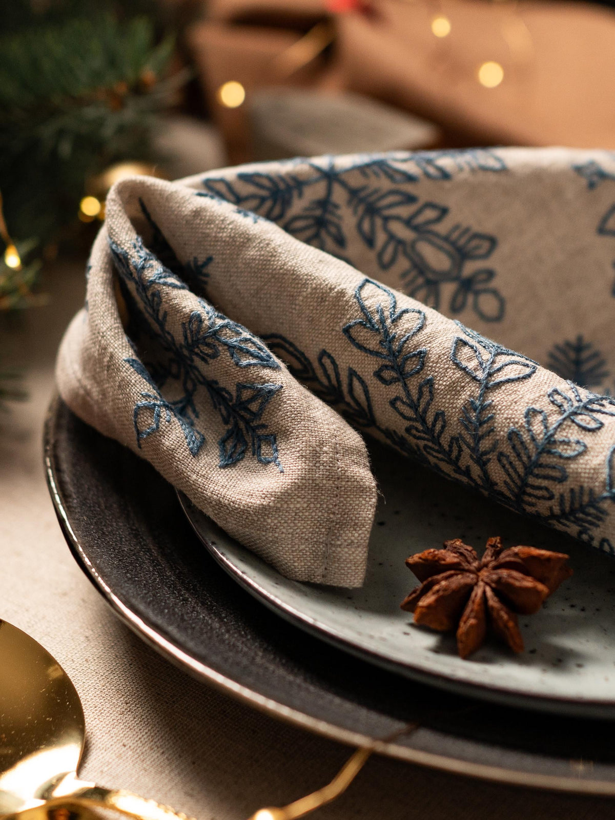 May include: A folded beige linen napkin with blue embroidered floral designs rests on a gray plate. A star anise sits on a light blue plate, with a gold spoon visible in the foreground. The scene suggests a festive dining setting.