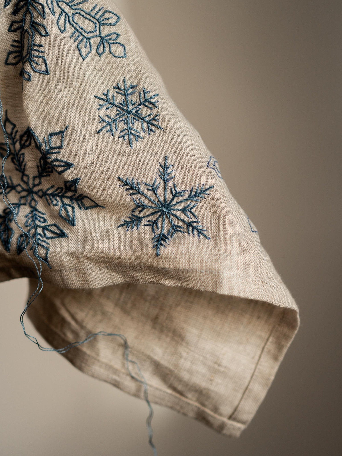 May include: Close-up of a beige fabric sleeve with embroidered blue snowflakes. The snowflakes vary in size and design, with intricate details. The fabric has a natural, textured appearance, and loose threads are visible.