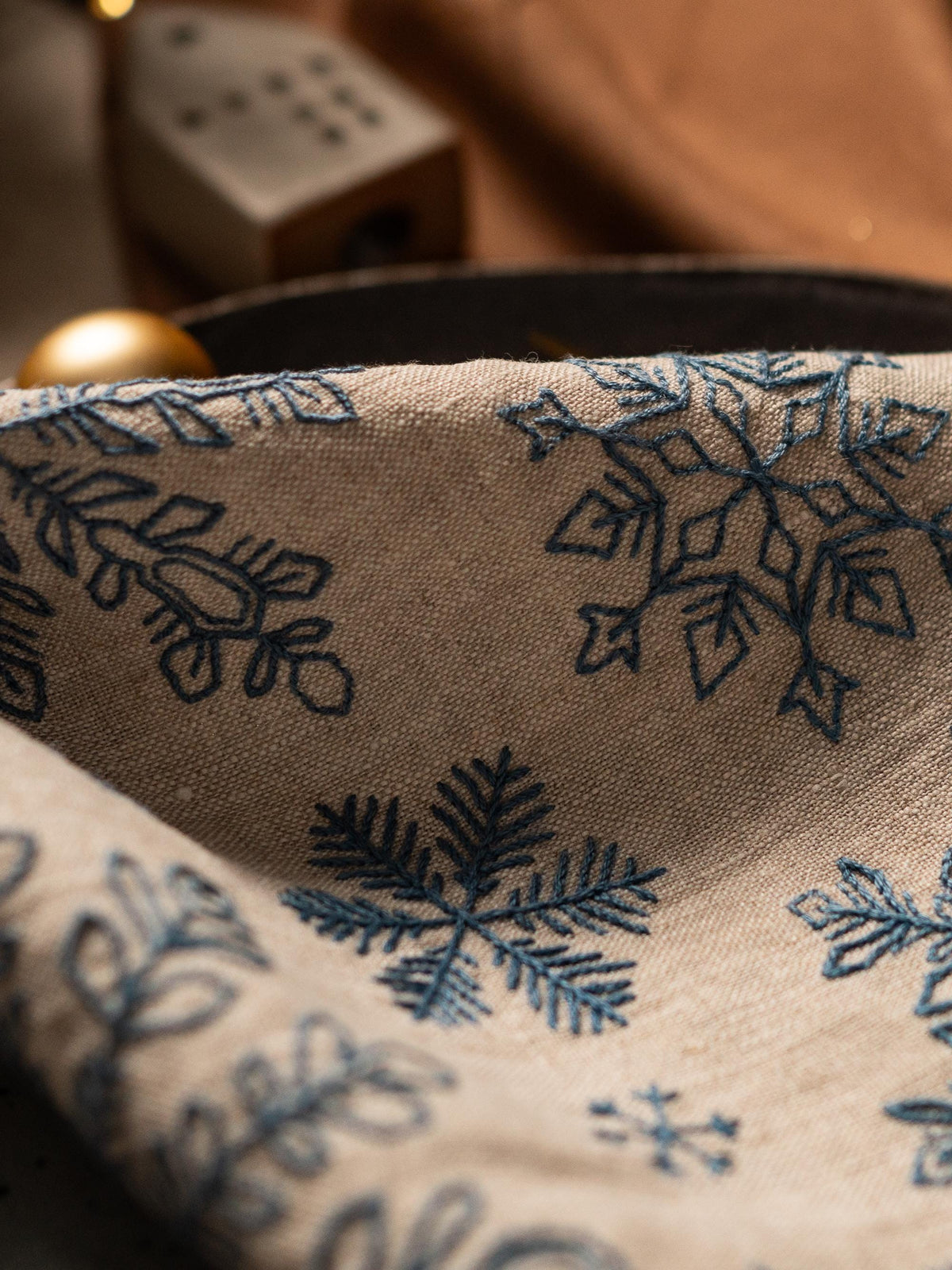 May include: Close-up of a beige linen fabric with embroidered blue snowflakes. The fabric is folded, revealing the intricate details of the winter-themed design. The image suggests a festive or seasonal product.