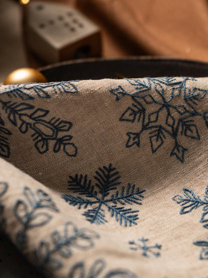 May include: Close-up of a beige linen fabric with embroidered blue snowflakes. The fabric is folded, revealing the intricate details of the winter-themed design. The image suggests a festive or seasonal product.