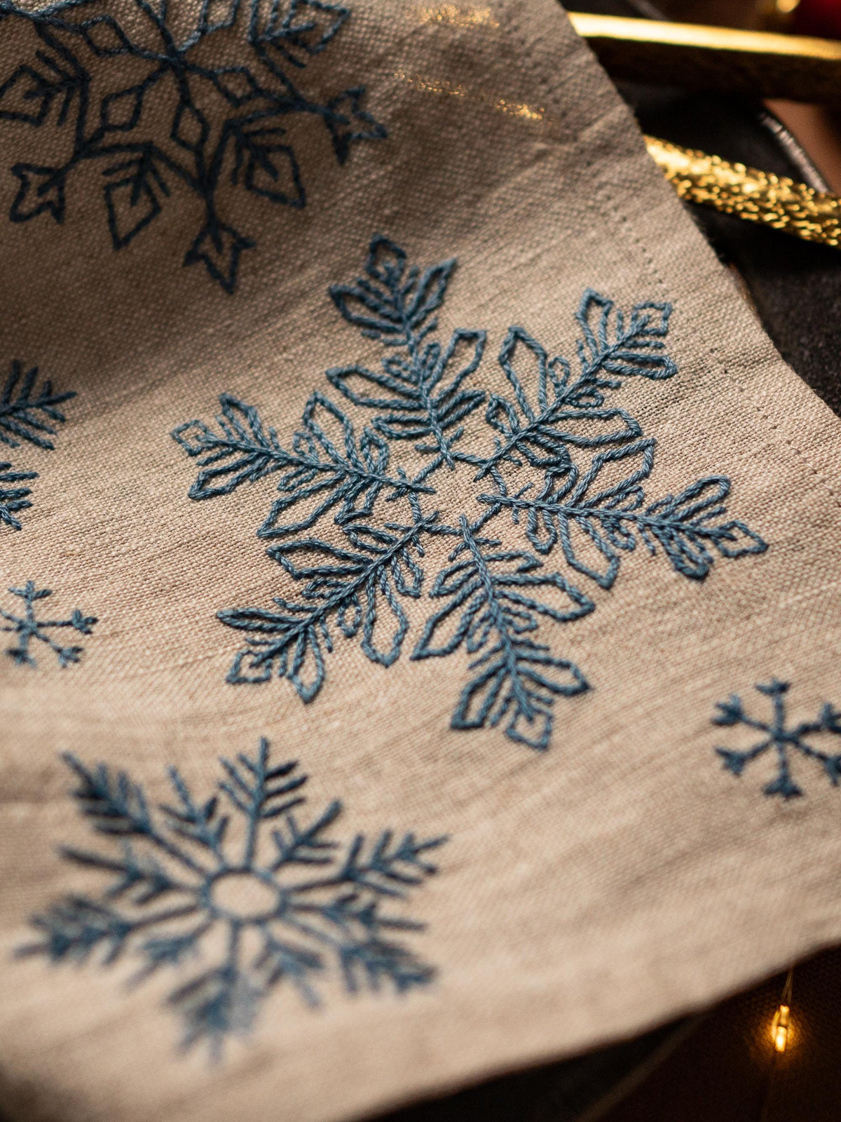 May include: Close-up of a beige linen fabric with embroidered blue snowflakes. The snowflakes vary in size and design, creating a wintery theme. The fabric has a textured appearance, suggesting a natural material.