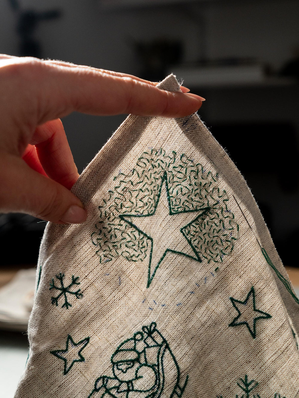 May include: Close-up of a beige linen fabric with green embroidered Christmas-themed designs. The designs include a star, snowflakes, and a Santa Claus figure. The fabric is being held by a hand.