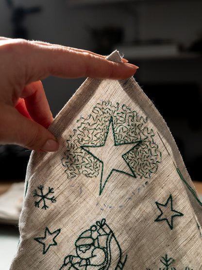 May include: Close-up of a beige linen fabric with green embroidered Christmas-themed designs. The designs include a star, snowflakes, and a Santa Claus figure. The fabric is being held by a hand.