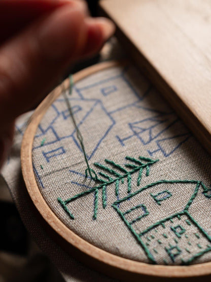 May include: Close-up of an embroidery project in progress. A wooden hoop holds gray fabric with a blue house design. Green thread is used to stitch a tree and outline the houses. A hand is visible, holding the needle.