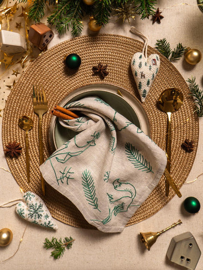 May include: A festive Christmas table setting with a woven placemat, gold utensils, and a linen napkin with green embroidered designs. Includes decorative ornaments, a bell, and cinnamon sticks.