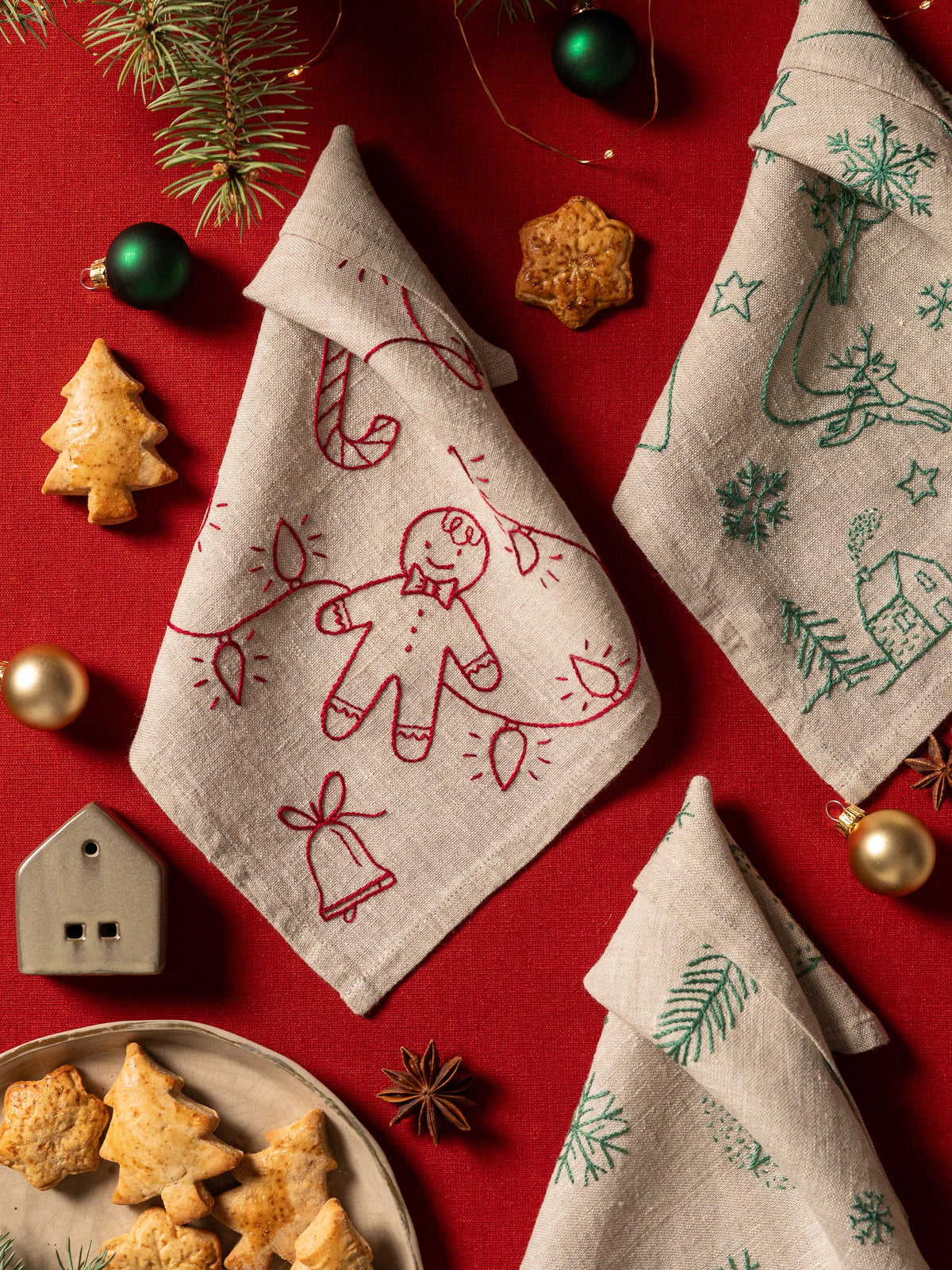 May include: A festive arrangement featuring folded linen napkins with embroidered Christmas designs. One napkin has a gingerbread man, candy cane, and lights. Another has a Christmas tree, reindeer, and house. The scene includes cookies, ornaments, 