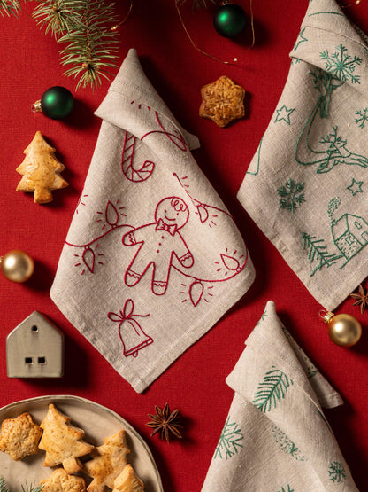 May include: A festive arrangement featuring folded linen napkins with embroidered Christmas designs. One napkin has a gingerbread man, candy cane, and lights. Another has a Christmas tree, reindeer, and house. The scene includes cookies, ornaments, 