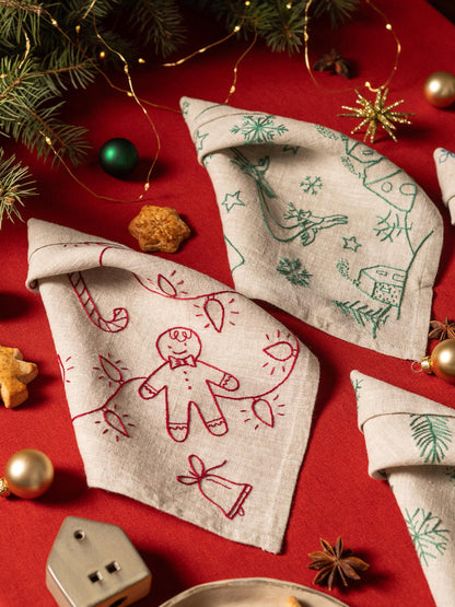 May include: Beige linen napkins with embroidered Christmas designs. One napkin features a gingerbread man and string lights in red, another has green snowflakes and a reindeer. Festive holiday decor.
