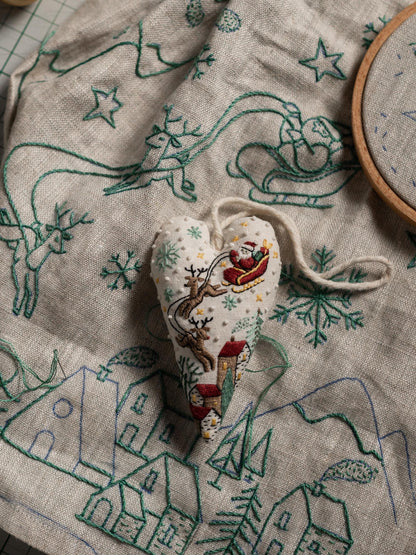 May include: A heart-shaped Christmas ornament with a Santa Claus design, reindeer, and houses. The ornament is white with embroidered details and a string for hanging. The background is a linen fabric with embroidered Christmas scenes.