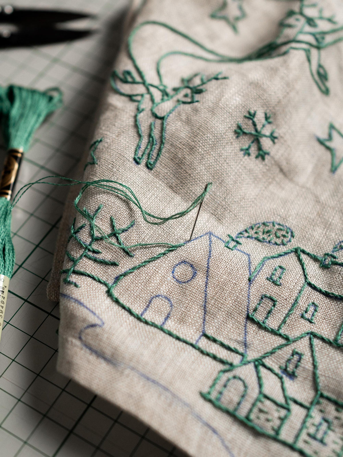 May include: Close-up of a beige fabric with green embroidery. The embroidery depicts a winter scene with houses, reindeer, snowflakes, and stars. A spool of green thread and a needle are visible, indicating a craft project.
