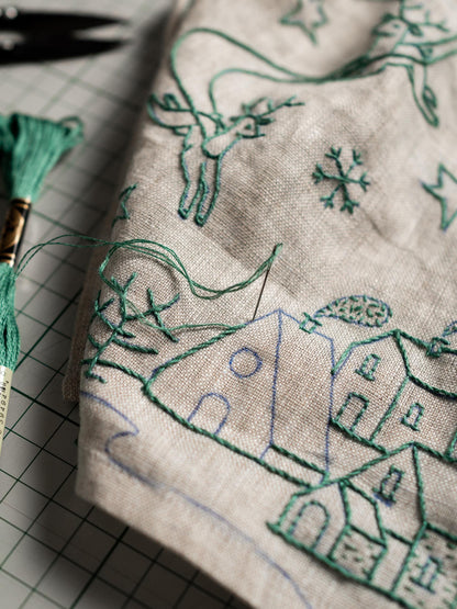 May include: Close-up of a beige fabric with green embroidery. The embroidery depicts a winter scene with houses, reindeer, snowflakes, and stars. A spool of green thread and a needle are visible, indicating a craft project.