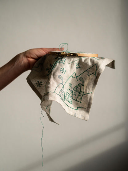May include: An embroidery hoop holds a linen fabric with a green stitched winter village scene. The design includes houses, mountains, snowflakes, and stars. The thread is still attached, indicating an in-progress embroidery project.