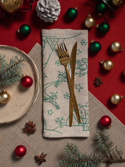 May include: A festive holiday table setting with a linen napkin featuring green embroidered winter scenes, gold cutlery, and red and gold Christmas ornaments. A plate with pine sprigs and star anise complete the arrangement.