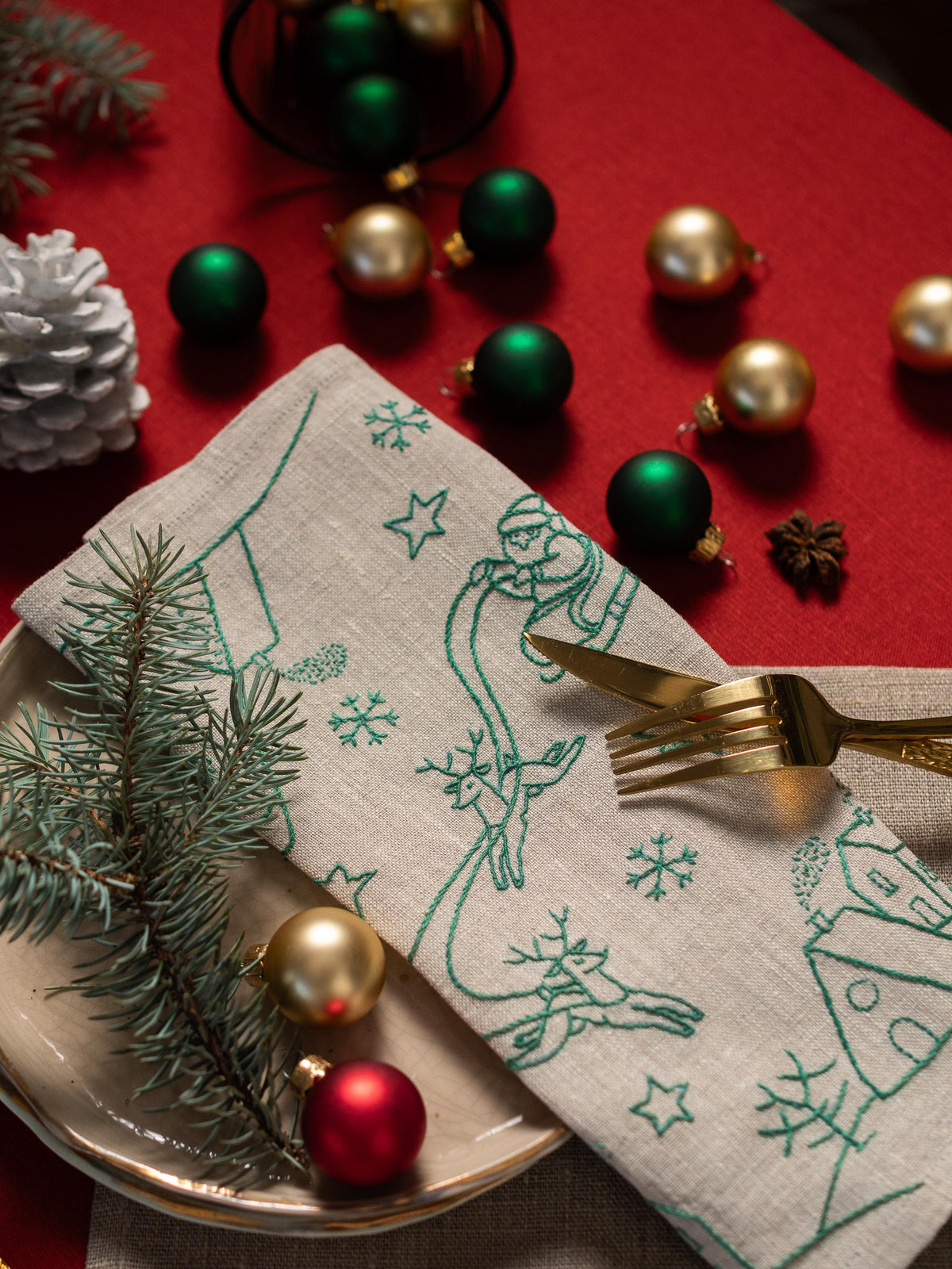 May include: A festive Christmas table setting with a linen napkin featuring green embroidered holiday designs, including Santa Claus and reindeer. Gold and green ornaments, a pine cone, and a sprig of evergreen are arranged on a red tablecloth.
