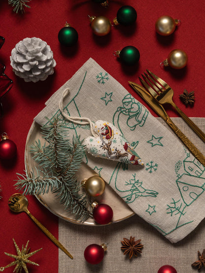May include: A festive Christmas table setting with a red tablecloth, featuring a plate with a linen napkin, gold cutlery, and ornaments. The napkin has green embroidered designs of snowflakes, stars, and reindeer. A heart-shaped ornament and pine sp