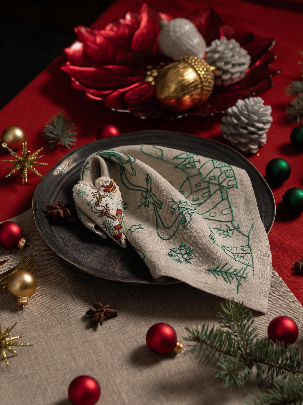 May include: A festive Christmas table setting with a linen napkin featuring a green embroidered winter village design. The scene includes red and gold ornaments, pine cones, and a heart-shaped napkin ring.