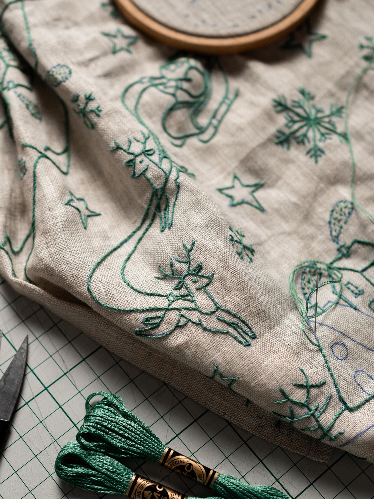 May include: Close-up of a linen fabric with green embroidered holiday designs, including reindeer, snowflakes, and stars. Two spools of green embroidery floss and a pair of scissors are in the foreground. A wooden embroidery hoop is visible.
