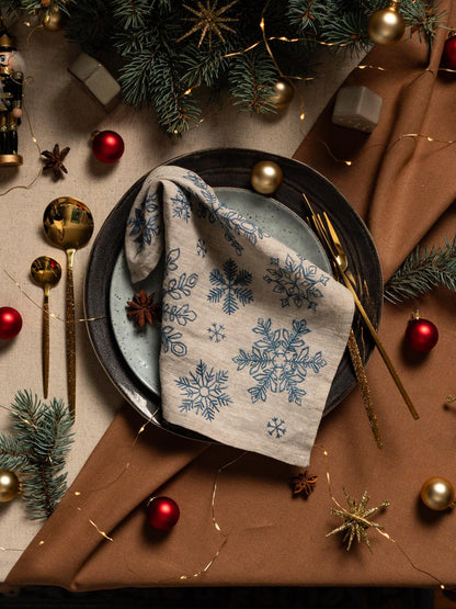 May include: A festive table setting with a beige linen napkin featuring blue snowflake embroidery. The plate is set with gold cutlery, red ornaments, and a sprig of evergreen. A gold star and string lights add to the holiday decor.