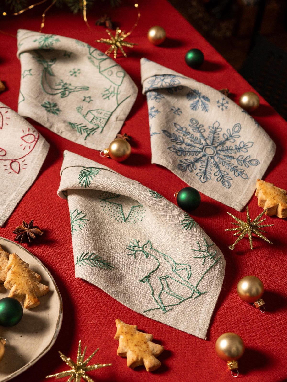 May include: A festive arrangement featuring folded linen napkins with embroidered holiday designs. The napkins, in natural tones, showcase green and blue snowflake, deer, and winter scene motifs. Gold and green ornaments and star-shaped decorations 