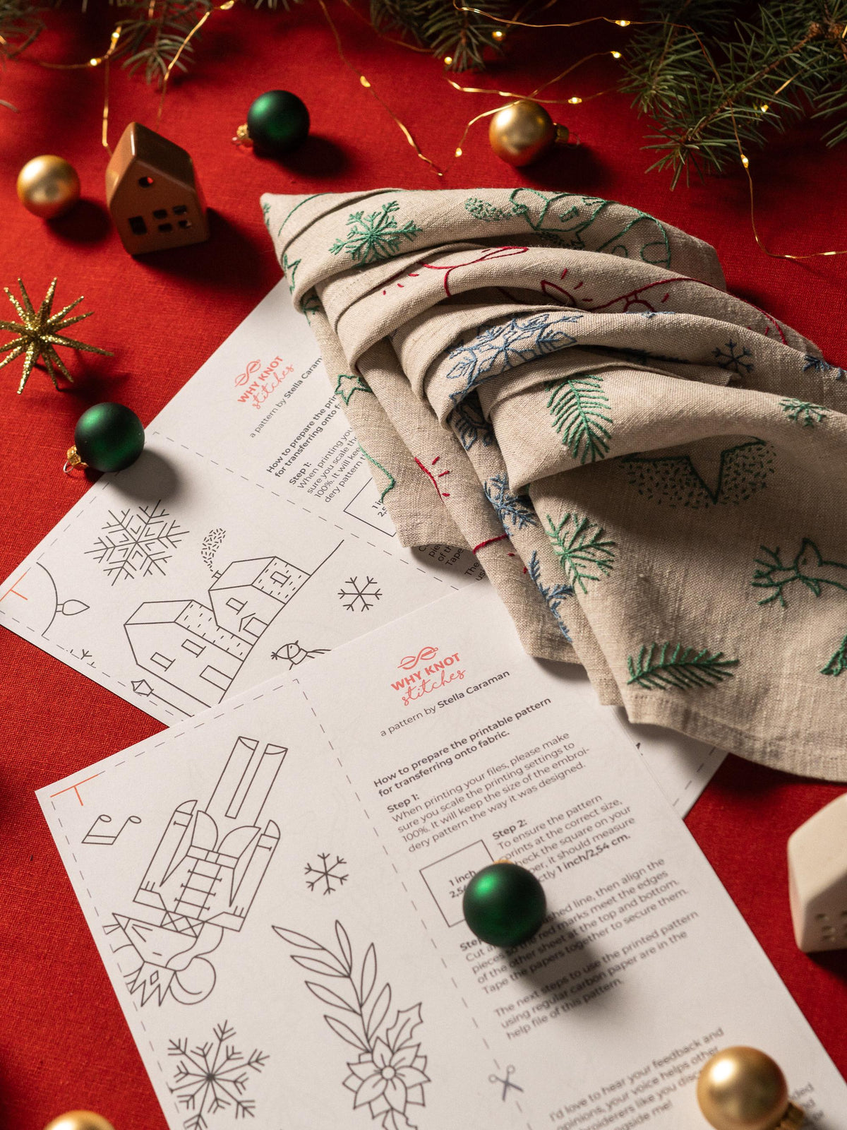 May include: A folded beige linen fabric with embroidered green, red, and blue Christmas designs, alongside printed patterns and Christmas ornaments on a red surface. The patterns include snowflakes and houses.