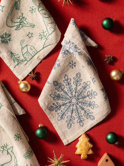 May include: Beige linen napkins with embroidered Christmas designs, including snowflakes, reindeer, and houses. The napkins are arranged on a red surface with green and gold ornaments, star anise, and a gingerbread tree.