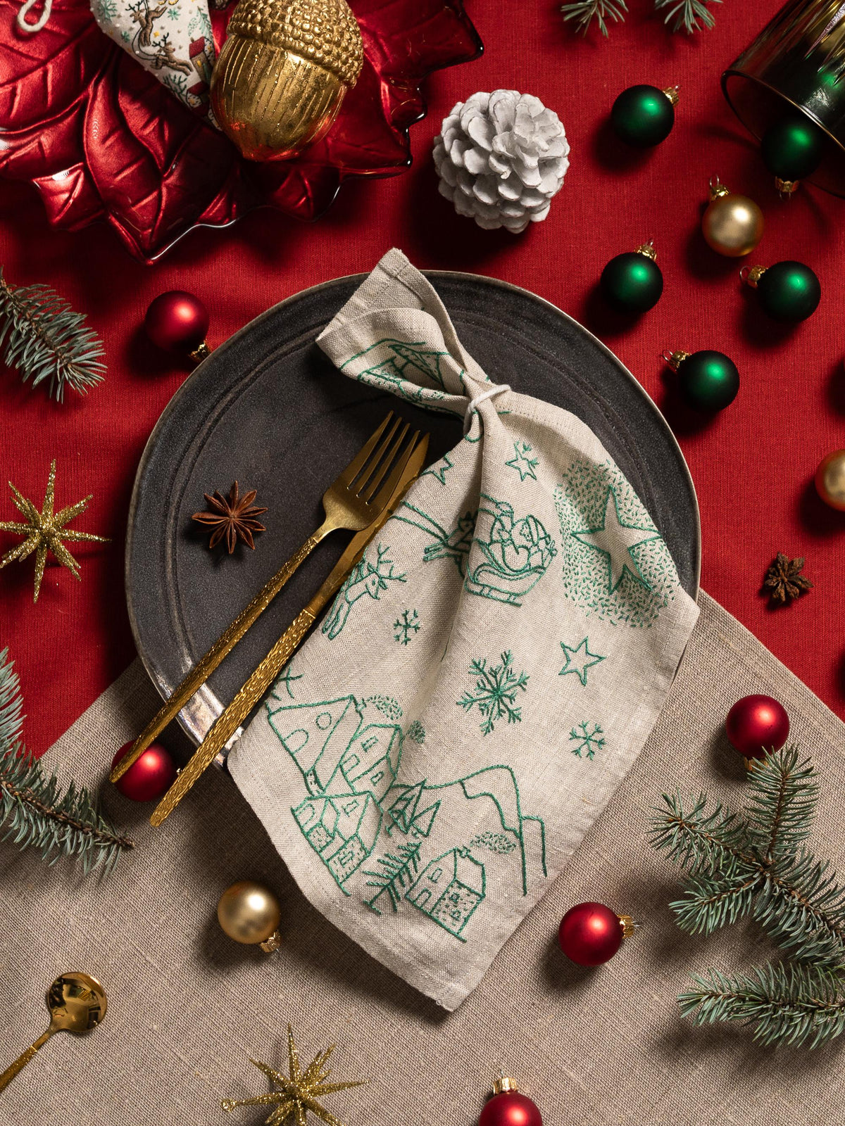 May include: A festive Christmas table setting with a gray plate, gold utensils, and a linen napkin embroidered with a green winter scene. Red and gold ornaments, pine needles, and star-shaped decorations complete the holiday theme.