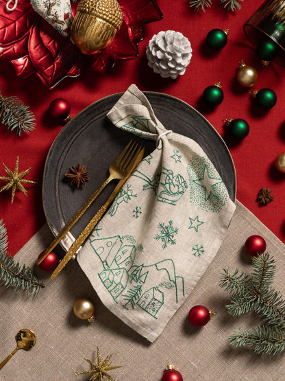 May include: A festive Christmas table setting with a gray plate, gold utensils, and a linen napkin embroidered with a green winter scene. Red and gold ornaments, pine needles, and star-shaped decorations complete the holiday theme.