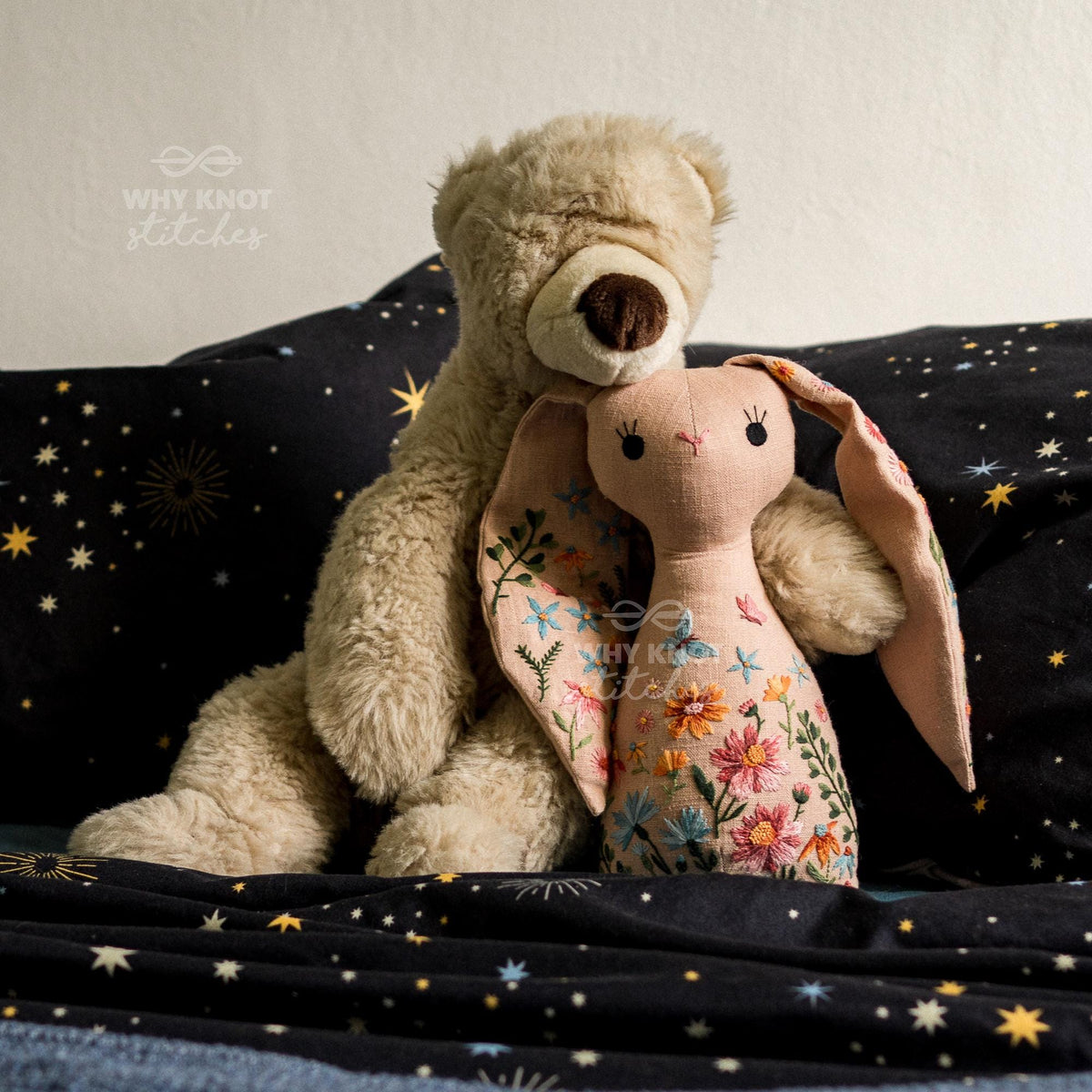 May include: A plush beige teddy bear hugging a light pink fabric rabbit doll. The rabbit features embroidered floral patterns in orange, pink, and blue on its body and ears, with simple black stitched eyes and a small nose. The text Why Knot Stitche
