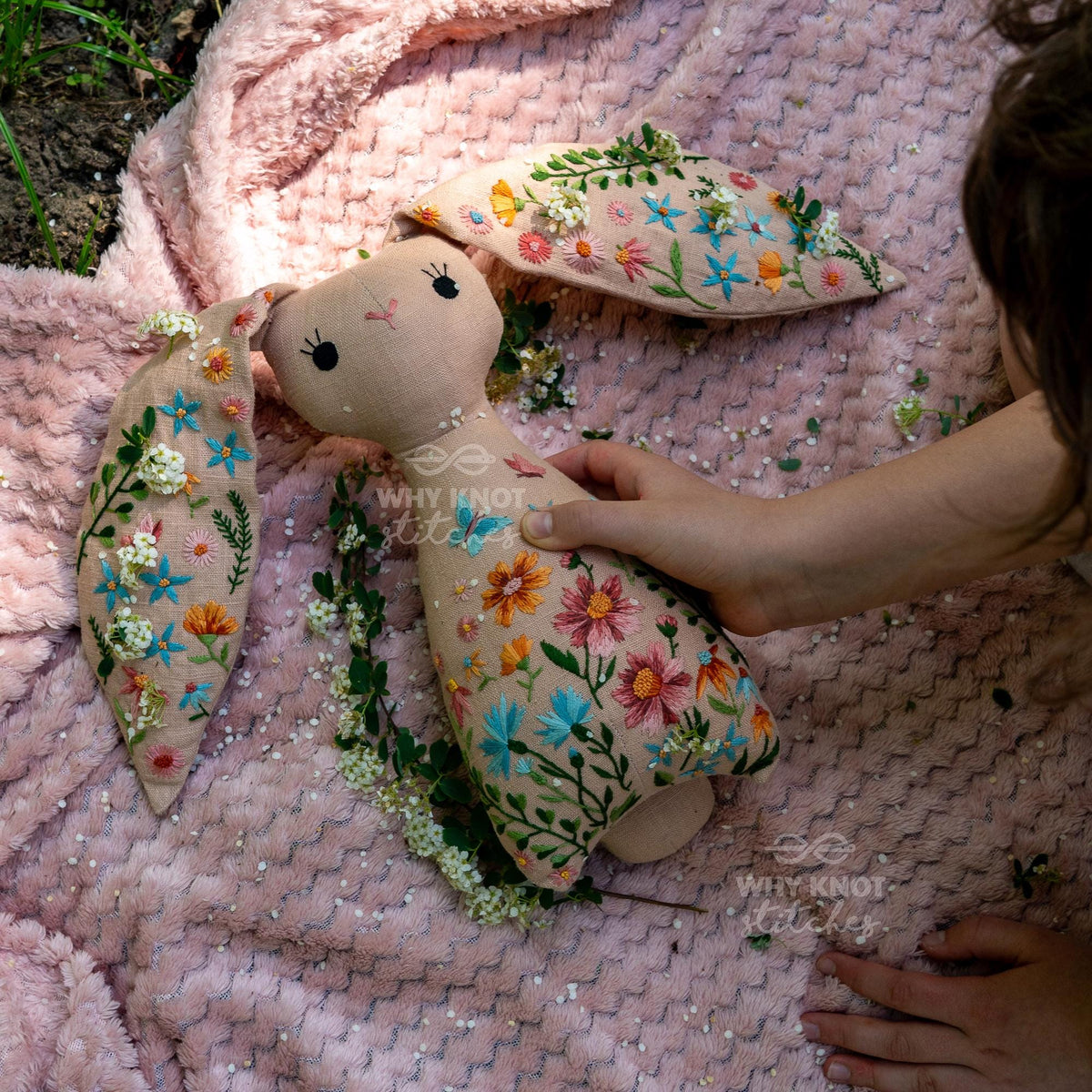 May include: A beige fabric rabbit doll featuring intricate floral embroidery in shades of pink, blue, orange, and green. The rabbit has long ears, a simple stitched face, and the text Why Knot Stitches is visible on the body and ear.
