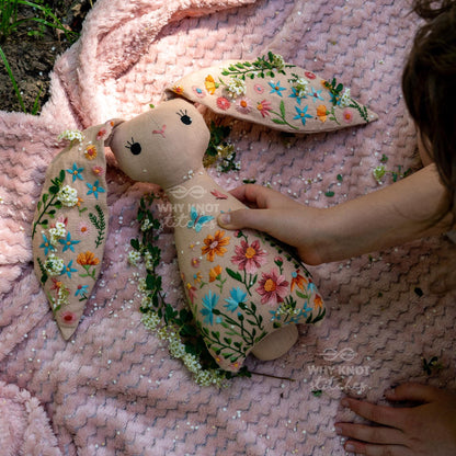 May include: A beige fabric rabbit doll featuring intricate floral embroidery in shades of pink, blue, orange, and green. The rabbit has long ears, a simple stitched face, and the text Why Knot Stitches is visible on the body and ear.