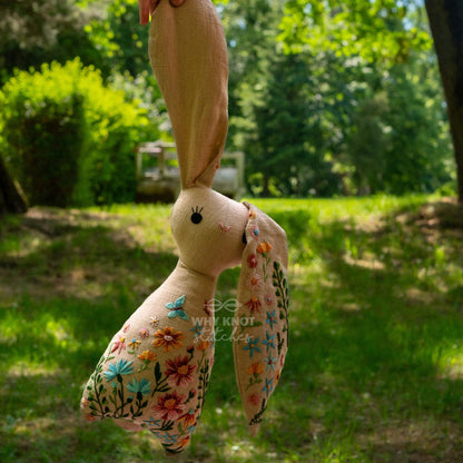 May include: A beige fabric rabbit toy featuring intricate floral embroidery in shades of blue, orange, pink, and green. The design includes a small blue butterfly and the text "WHY KNOT STITCHES" embroidered on the body. It has long ears.