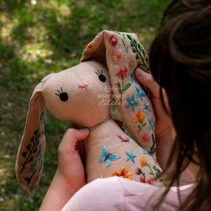 May include: A beige fabric rabbit doll featuring embroidered floral and butterfly motifs in shades of blue, orange, pink, and green. The rabbit has black stitched eyes and a pink nose. The text "WHY KNOT STITCHES" is embroidered on the rabbit's head