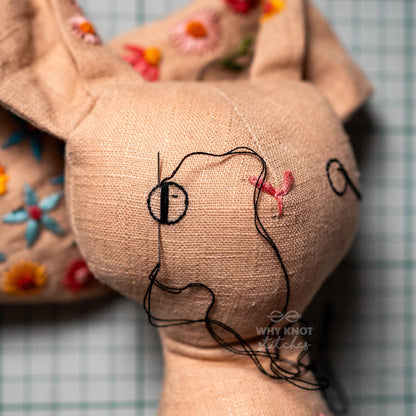 May include: A beige fabric stuffed animal with embroidered floral patterns in red, orange, and blue. A silver needle is inserted into the head, creating a black circular eye with black thread trailing down. The text Why Knot Stitches is visible.