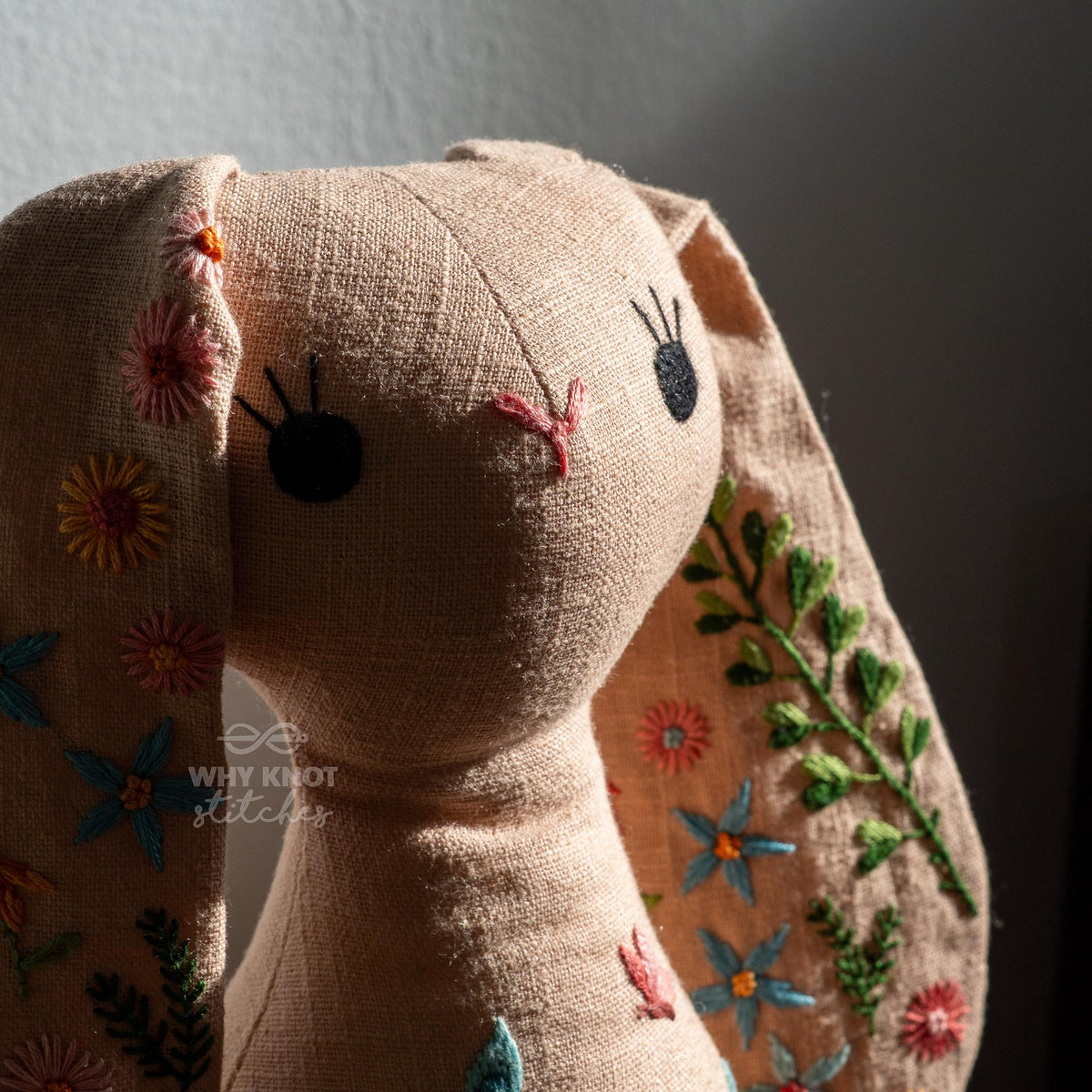 May include: A beige fabric rabbit doll featuring embroidered floral patterns in pink, orange, blue, and green on its long ears. The face has black stitched eyes with eyelashes and a pink stitched nose. The text Why Knot Stitches is visible.