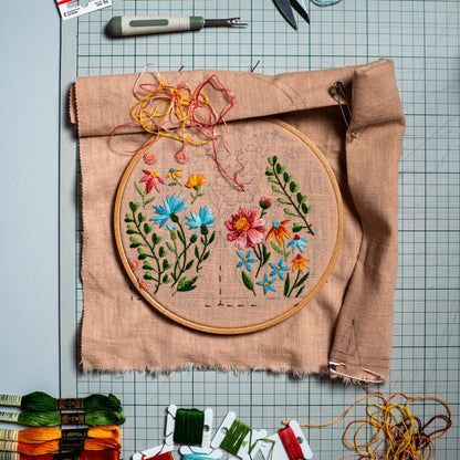 May include: A round wooden embroidery hoop holding light brown fabric with a partially finished floral design featuring blue, pink, orange, and green thread. Loose colorful embroidery floss, a seam ripper, and thread bobbins are arranged nearby.