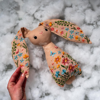 May include: A beige fabric rabbit plush toy featuring intricate floral embroidery in shades of pink, orange, blue, and green. The rabbit has long ears, black stitched eyes, and a pink nose. The text "WHY KNOT STITCHES" is visible on the chest.
