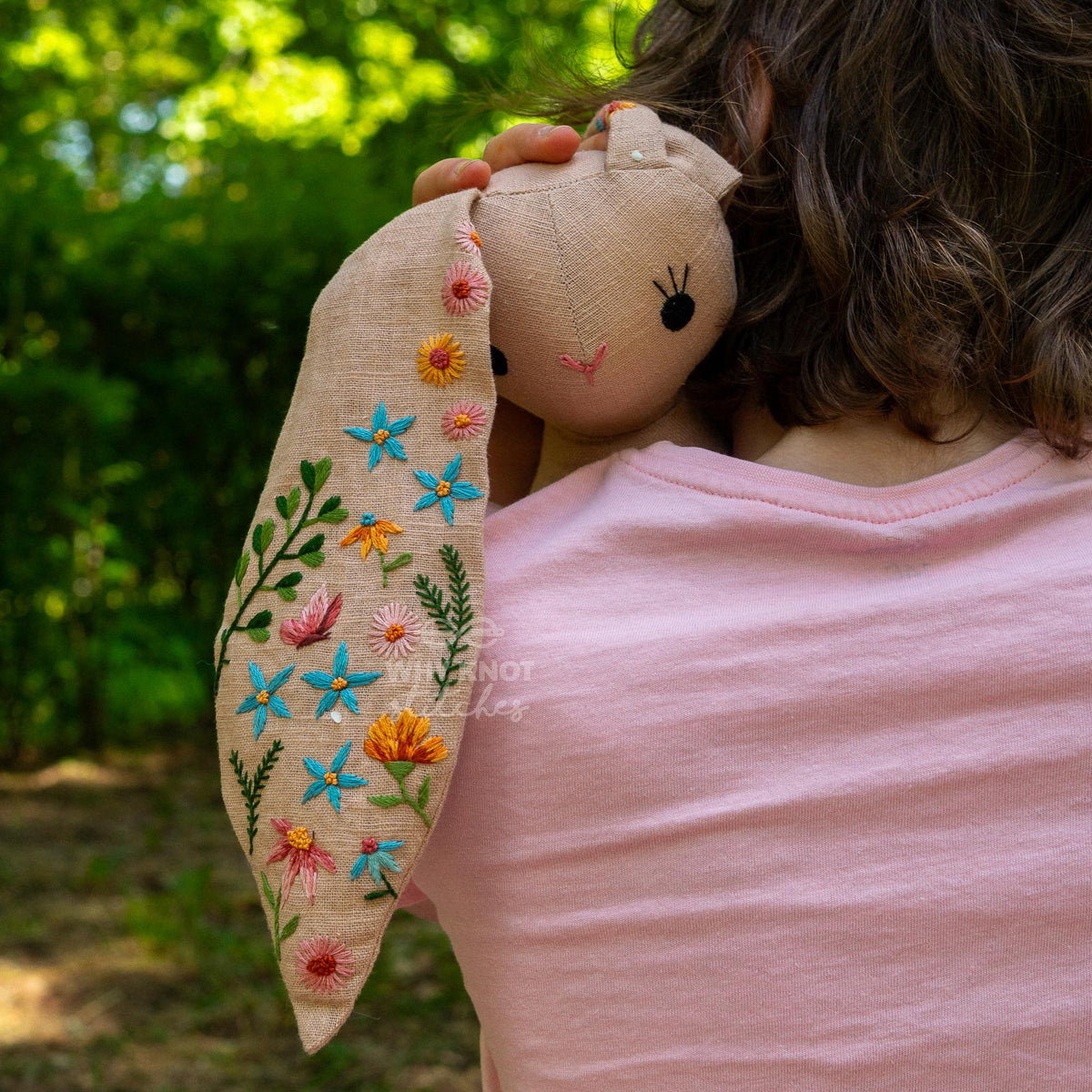 May include: A beige linen rabbit doll featuring long ears embroidered with colorful floral patterns including blue, orange, pink, and green thread. The rabbit has simple black stitched eyes and a small pink stitched nose, held against a light pink s