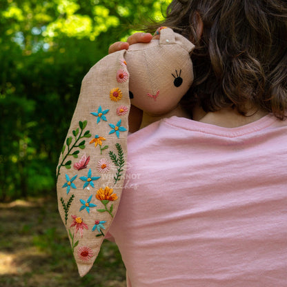 May include: A beige linen rabbit doll featuring long ears embroidered with colorful floral patterns including blue, orange, pink, and green thread. The rabbit has simple black stitched eyes and a small pink stitched nose, held against a light pink s