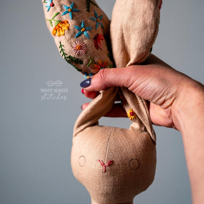 May include: A tan linen stuffed rabbit toy with long ears featuring colorful floral embroidery in blue, orange, and green. The face has simple stitched circular eyes and a pink nose. The text Why Knot Stitches is printed in white on the left side.