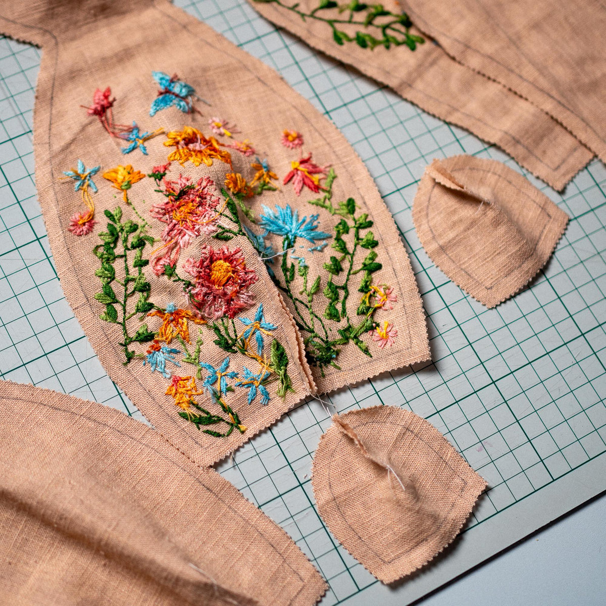 May include: Several pieces of light brown linen fabric featuring intricate floral embroidery in vibrant shades of pink, yellow, orange, and blue. The fabric pieces are cut into various shapes, including a bodice-like panel and small rounded sections