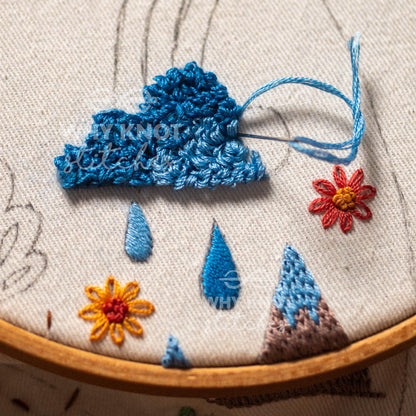 May include: An embroidery hoop containing a fabric panel with a three-dimensional crocheted blue cloud, two embroidered blue raindrops, a brown mountain peak, and two small flowers in red and yellow, with a needle and blue thread attached.