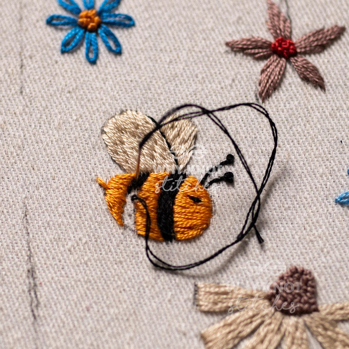 May include: An embroidery piece on light beige fabric featuring a central bee with yellow and black stripes, light tan wings, and two antennae. Surrounding the bee are embroidered flowers in blue and brown, with a loose black thread loop.