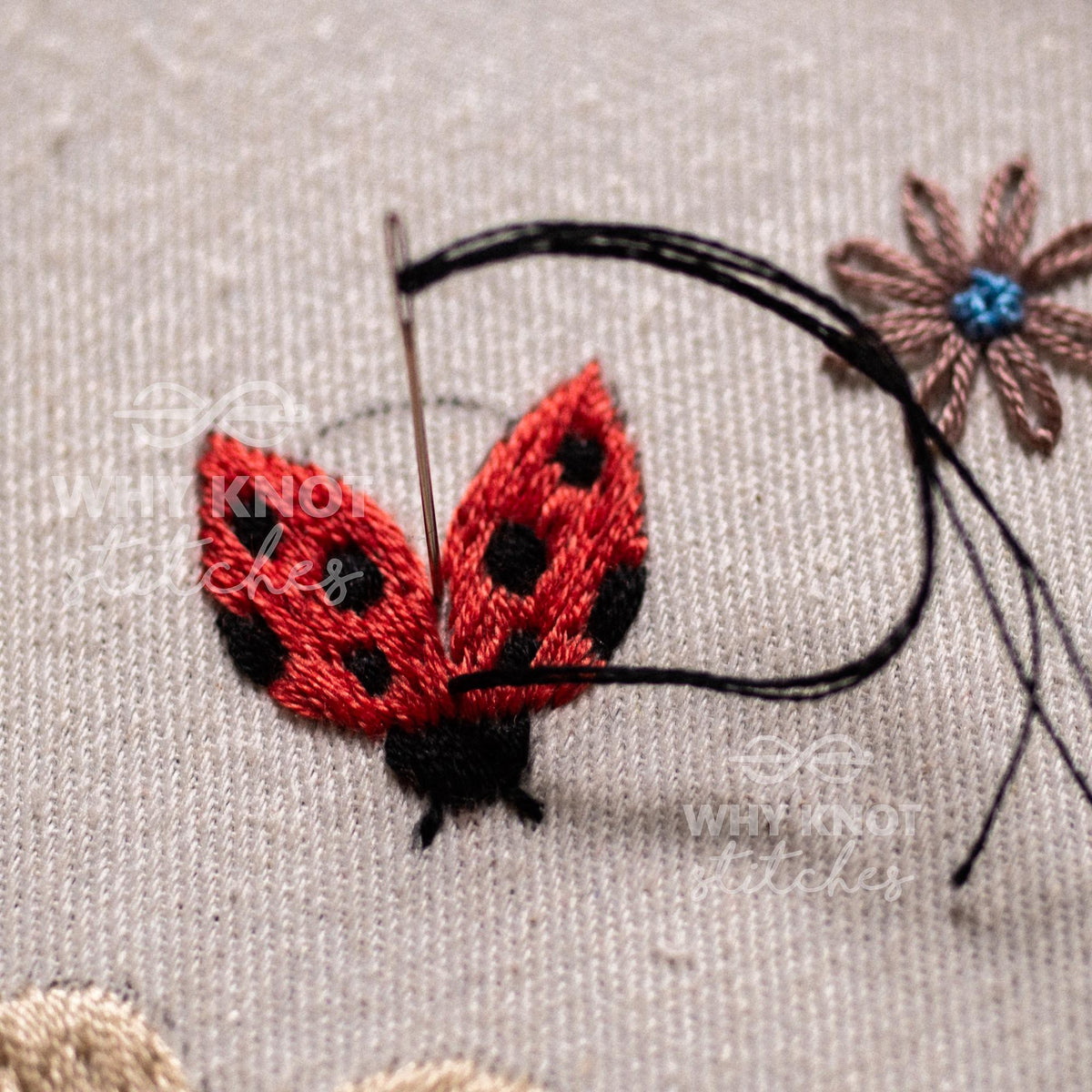 May include: An embroidery project on light beige fabric featuring a red ladybug with black spots, stitched with a needle and black thread. A small brown flower with a blue center is embroidered to the right of the ladybug on the fabric.